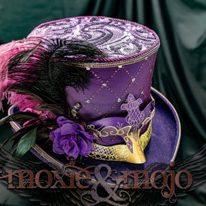 May include: A purple top hat featuring a diamond-patterned crown with silver sequins, a wide brim, and a decorative mask attached to the front. The hat is adorned with black and pink ostrich feathers and a purple fabric rose. Text reads moxie & mojo.