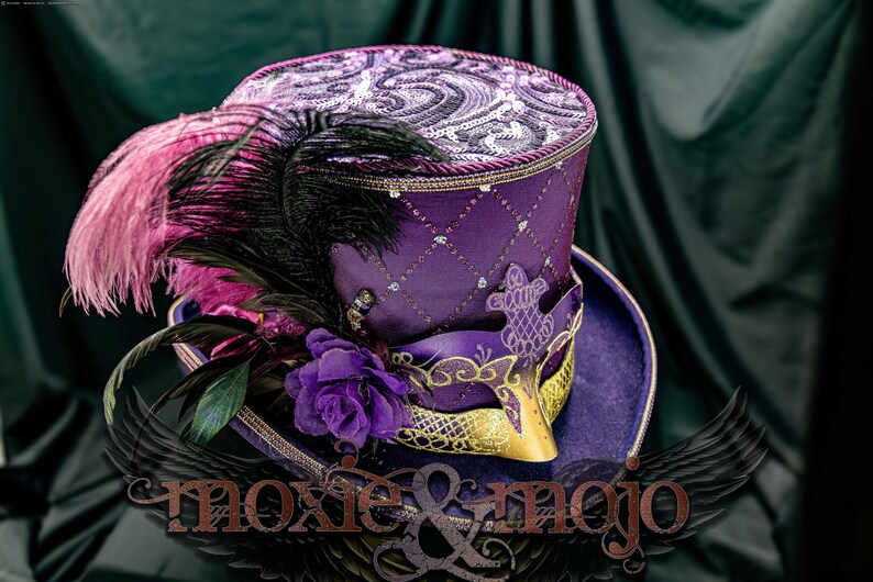May include: A purple top hat featuring a diamond-patterned crown with silver sequins, a wide brim, and a decorative mask attached to the front. The hat is adorned with black and pink ostrich feathers and a purple fabric rose. Text reads moxie & mojo.