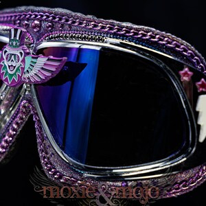 Puede incluir: Gafas protectoras con purpurina morada que presentan un emblema central de oso alado con sombrero de copa, lentes oscuras reflectantes y un rayo en el lateral. El marco est&aacute; decorado con cadenas moradas met&aacute;licas y peque&ntilde;as estrellas rosas.