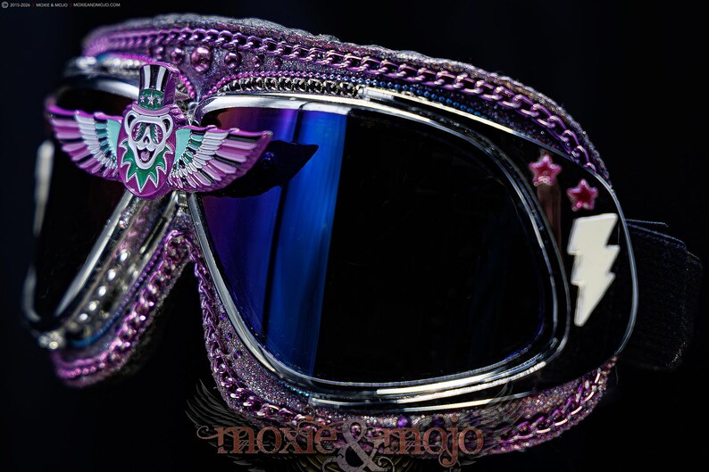 Puede incluir: Gafas protectoras con purpurina morada que presentan un emblema central de oso alado con sombrero de copa, lentes oscuras reflectantes y un rayo en el lateral. El marco est&aacute; decorado con cadenas moradas met&aacute;licas y peque&ntilde;as estrellas rosas.