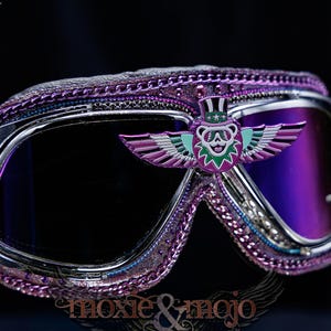 Puede incluir: Gafas protectoras moradas con montura de metal plateado y lentes de color morado oscuro. La montura est&aacute; decorada con un borde de cadena morada y un emblema central de un oso alado con sombrero de copa. Incluye detalles de estrellas y rayos.