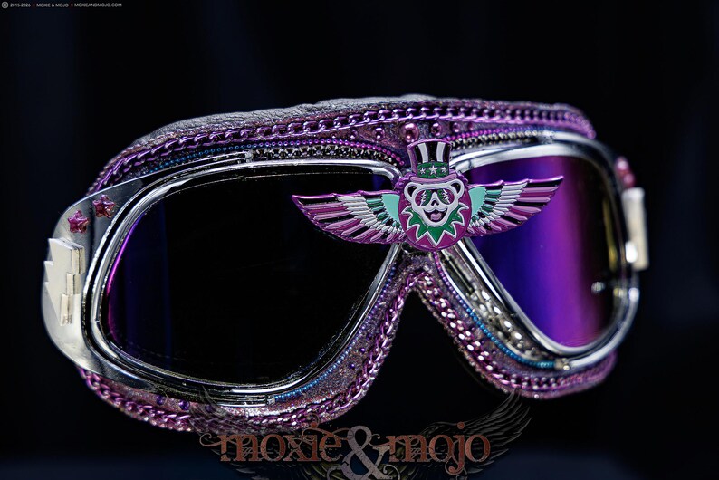 Puede incluir: Gafas protectoras moradas con montura de metal plateado y lentes de color morado oscuro. La montura est&aacute; decorada con un borde de cadena morada y un emblema central de un oso alado con sombrero de copa. Incluye detalles de estrellas y rayos.