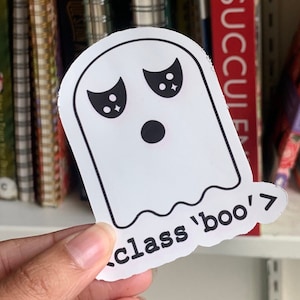 May include: A white die-cut sticker featuring a black outline of a ghost with large, expressive eyes and a round mouth. The text 'class 'boo'>' is printed in black at the bottom of the sticker, which has a smooth, matte-like finish and rounded edges.