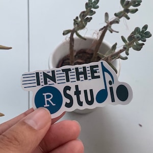 May include: A die-cut vinyl sticker featuring the text IN THE R Studio in black and blue lettering. The letter R is inside a blue circle, and the letter o is replaced by a blue musical note symbol. Horizontal blue lines extend from the text.