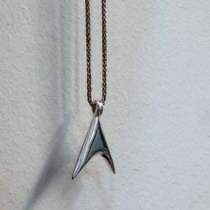 May include: A silver-colored pendant necklace with a triangular design. The pendant is suspended from a delicate chain. The pendant has a polished, reflective surface, and the chain is a woven style.