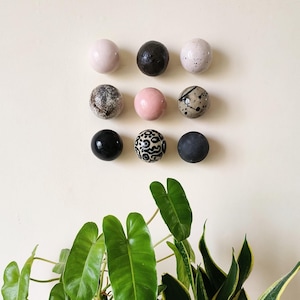 Könnte beinhalten: Neun runde Keramik-Ornamente in verschiedenen Farben und Mustern, darunter Schwarz, Weiß, Rosa und Braun. Die Ornamente sind in einem Kreis an einer weißen Wand angeordnet.