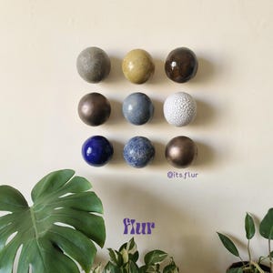 Könnte beinhalten: Neun dekorative Kugeln in verschiedenen Farben und Texturen sind an einer cremefarbenen Wand angeordnet. Die Kugeln umfassen Grautöne, Braun, Blau und Weiß mit glatten, strukturierten und gesprenkelten Oberflächen. Das Wort "flur" ist in lila Schrift geschrieben.