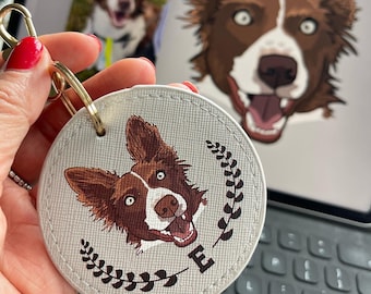 May include: A white leather keychain with a brown and white border collie dog illustration. The dog is smiling with its tongue out. The keychain has a gold metal ring and a white background with a brown floral design.