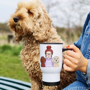Può includere: Tazza da viaggio bianca con un'illustrazione a cartoni animati di una donna, occhiali e due cani, con la scritta "Dogs Before Dudes" su una maglietta viola. La tazza ha un manico e una base neri. Sullo sfondo c'è un cane marrone chiaro e soffice.