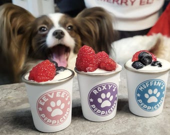 May include: Three small white cups with silver rims, each topped with cream and berries. The cups have different colored paw print logos and text: "Evee's Puppacino", "Roxy's Puppacino", and "Muffin's Puppacino". A dog is in the background.