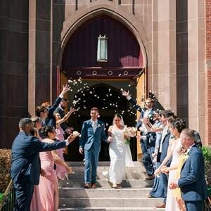 以下が含まれることがあります： 教会の入り口の石段を降りる結婚式のカップルと、白い花びらを投げる正装したゲストたち。カップルは中央に位置し、左側のパートナーは青いスーツ、右側のパートナーは白いドレスを着用している。