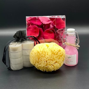 Puede incluir: Un set de regalo de spa con una esponja vegetal amarilla, una botella de vidrio transparente con un líquido rosa y una etiqueta blanca que dice "Rose Bubble Bath 2 oz", una bolsa de pétalos de rosa rosas y tres velas votivas blancas.