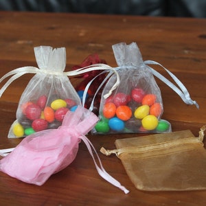 May include: Four small organza bags filled with colorful candy. The bags are white, pink, gold, and light blue. The bags are tied with ribbon.