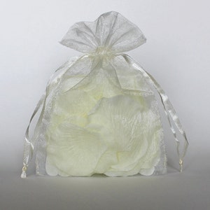 Puede incluir: Una bolsa de organza blanca transparente llena de pétalos de rosa marfil. La bolsa está atada con una cinta blanca.