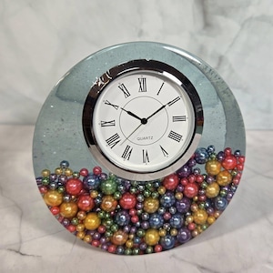 May include: A decorative quartz clock with a white face, black hands, and Roman numerals. The clock is encased in a clear, light blue resin with colorful beads at the base. The clock is approximately 5 inches in diameter.