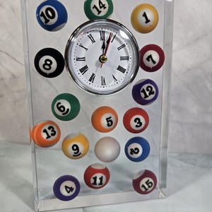 May include: A clear acrylic clock featuring a white clock face with black Roman numerals and a silver bezel. Billiard balls with numbers 1-15 are embedded in the acrylic. The clock is a unique decorative item.