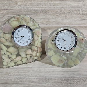 May include: Two quartz clocks encased in clear resin filled with off-white and pale pink stone fragments. One clock is arch-shaped, and the other is circular. Both feature white faces with black numerals, silver bezels, and black hour and minute hands.
