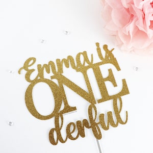 May include: Gold glitter cake topper with the text "Emma is One derful".