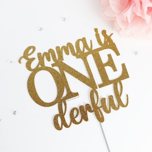 May include: Gold glitter cake topper with the text "Emma is one derful".