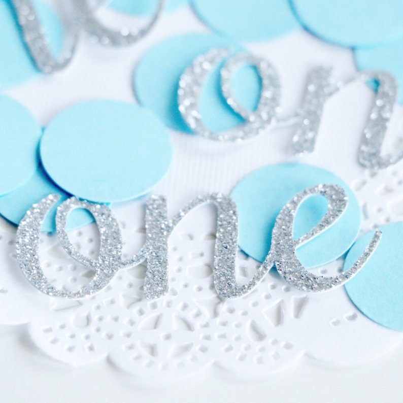 May include: Silver glitter "one" banner with blue confetti on a white lace background.