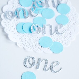 May include: Silver glitter 'one' cutouts with blue paper circles on a white doily.