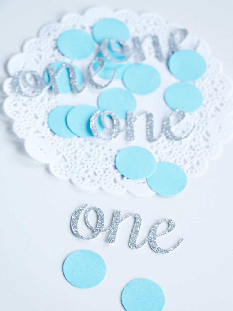 May include: Silver glitter 'one' cutouts with blue paper circles on a white doily.