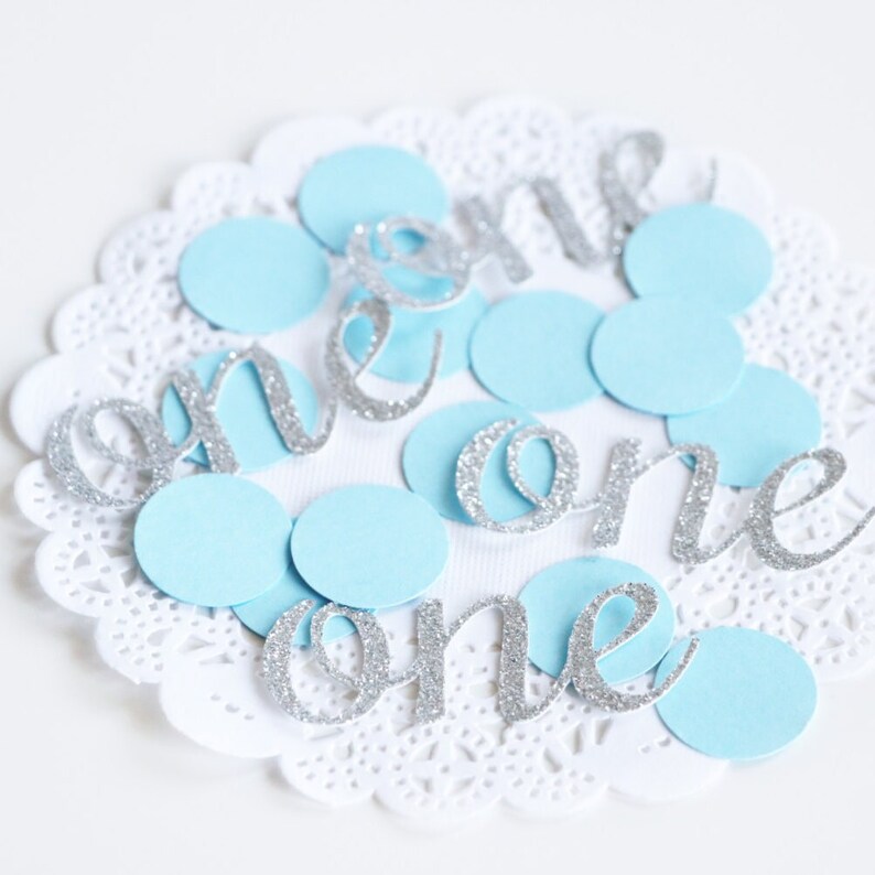 May include: Silver glitter "one" cutouts on a white doily with light blue confetti circles.