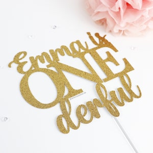 May include: Gold glitter cake topper with the name "Emma" and the word "one" in large letters, followed by the word "derful".