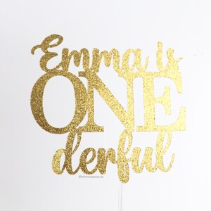 May include: Gold glitter cake topper with the text "Emma is one derful" and the social media handle "@athenasatelier.de".