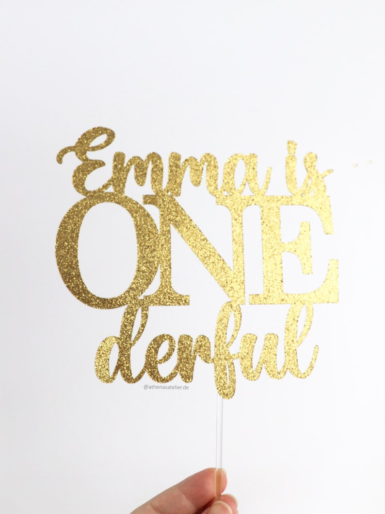 May include: Gold glitter cake topper with the text "Emma is one derful" and the social media handle "@athenasatelier.de".