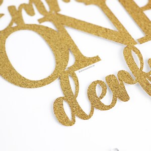 May include: Gold glitter cake topper with the words "Happy First Birthday" in a cursive font.