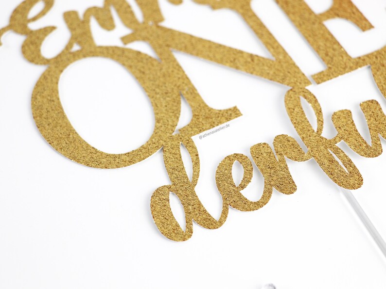 May include: Gold glitter cake topper with the words "Happy First Birthday" in a cursive font.