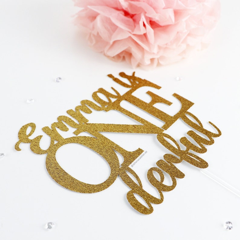 May include: Gold glitter cake topper with the text "Emma is One" in a cursive font.