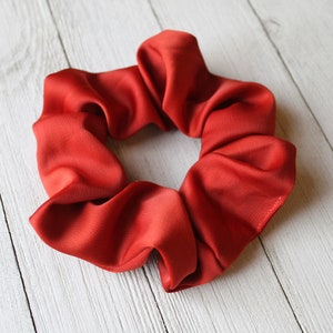 May include: A red scrunchie made of soft fabric. The scrunchie is in the shape of a circle and is lying flat on a white wooden surface.