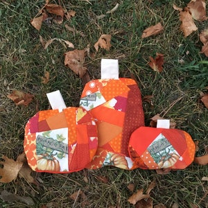Könnte beinhalten: Drei handgefertigte Stoffkürbisse in verschiedenen Größen, angeordnet auf Gras mit Herbstlaub. Die Kürbisse sind im Patchwork-Stil gehalten und weisen orangefarbene und rote Stoffe auf. Ein Kürbis trägt die Aufschrift "Pumpkin Farm".