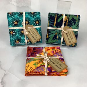 May include: Set of four fabric coasters with different colorful patterns. The coasters are tied together with a white ribbon and a brown tag that reads "Purse Your Lips Handmade Bags & More Coaster set (4)".
