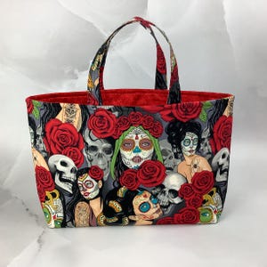 May include: A tote bag with a Day of the Dead theme. The bag features a gray background with sugar skulls, red roses, and women with painted faces. The interior is red, and the handles are black and gray.