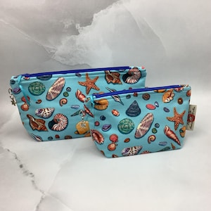May include: Two light blue zippered pouches featuring a repeating pattern of various seashells and orange starfish. The larger pouch has a blue zipper and a small metal charm, while the smaller pouch also features a blue zipper and a small tag.