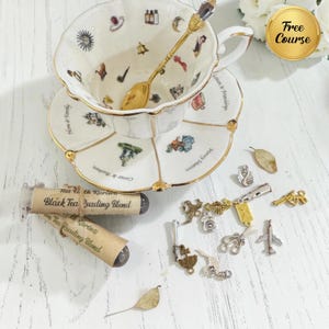 May include: A white porcelain teacup and saucer set with gold trim, featuring small colourful illustrations and text. A gold spoon rests inside the cup. Two tubes labelled Black Tea Reading Blend and various small metal charms are arranged nearby.