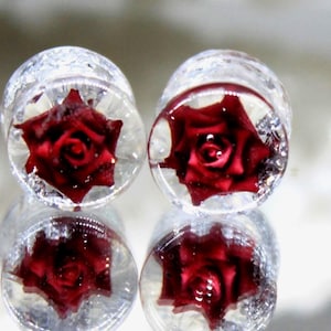 Puede incluir: Tapones para los oídos redondos y transparentes con un diseño de rosa roja. Las rosas están encapsuladas en resina transparente y los tapones tienen un aspecto brillante y con purpurina. Los tapones se reflejan en una superficie espejada.
