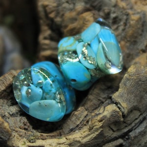 May include: Two turquoise blue glass plugs with gold flecks and a marbled pattern. The plugs are on a rough, brown wooden surface.