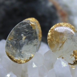 May include: Two clear teardrop-shaped resin earrings with gold leaf accents. The earrings are displayed on a bed of white quartz crystals.