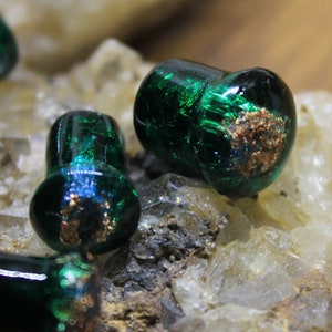 May include: Four green glass plugs with gold flecks, displayed on a bed of white crystals.