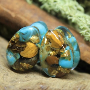 May include: A pair of teardrop-shaped resin ear gauges with embedded turquoise, tiger's eye, and gold flakes.