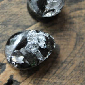 May include: Two black and silver gemstone plugs. The plugs are round and have a smooth, polished surface. They are sitting on a piece of wood.