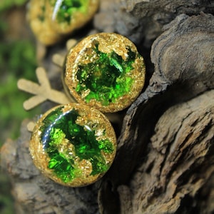 May include: Four round green and gold resin cabochons with gold leaf accents. The cabochons are set on a wooden surface.