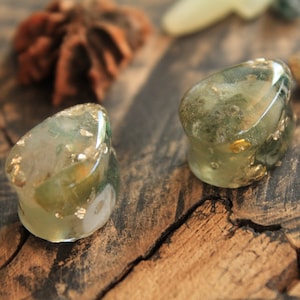 May include: Two green and gold resin ear gauges with a speckled pattern. The gauges are on a wooden surface.