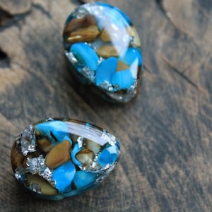 May include: Two teardrop-shaped resin earrings. Each earring features a mix of blue, brown, and silver elements. The blue stones are the most prominent, with brown and silver accents. The earrings are set against a dark wooden surface.