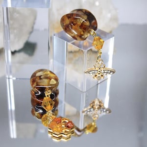 May include: A pair of amber-colored resin earrings with gold-colored bee charms. The earrings are displayed on clear acrylic blocks, reflecting on a mirrored surface. The bee charms are detailed with small crystals.