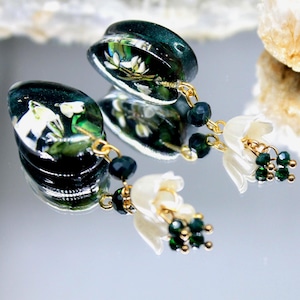 Puede incluir: Un par de pendientes florales verdes y blancos con cadenas doradas y cuentas verdes. Los pendientes presentan un colgante en forma de lágrima con un diseño floral y un colgante de flor blanca más pequeño.